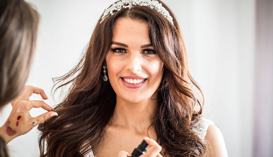 Porträt der lächelnden Braut mit Perlen-Diadem, während ihr die Haare gerichtet werden.