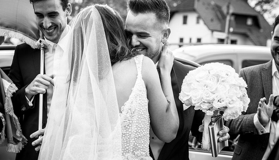 Der Bräutigam umarmt lachend die Braut vor dem Hochzeitsauto, Gäste und Häuser im Hintergrund.