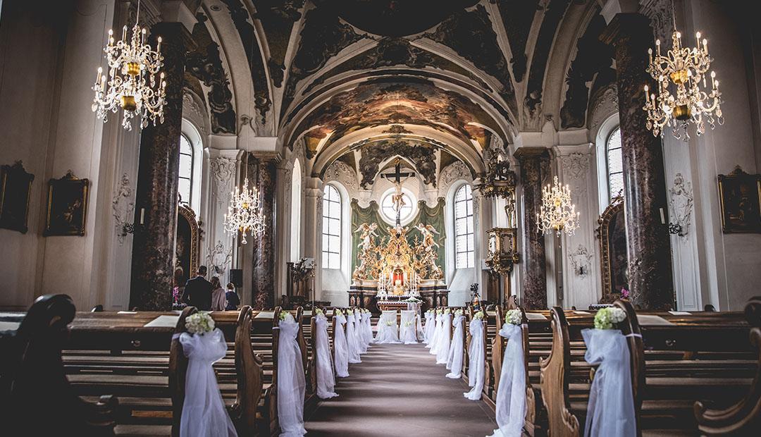 Blick in eine barocke Kirche mit Deckenfresken, zwei Kronleuchtern und weiß geschmückten Bankreihen zum Altar.