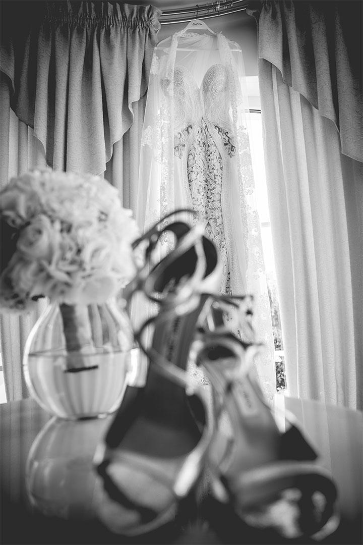 Brautkleid hängt am Fenster, davor Schuhe und ein Strauß weißer Blumen in einer Vase.