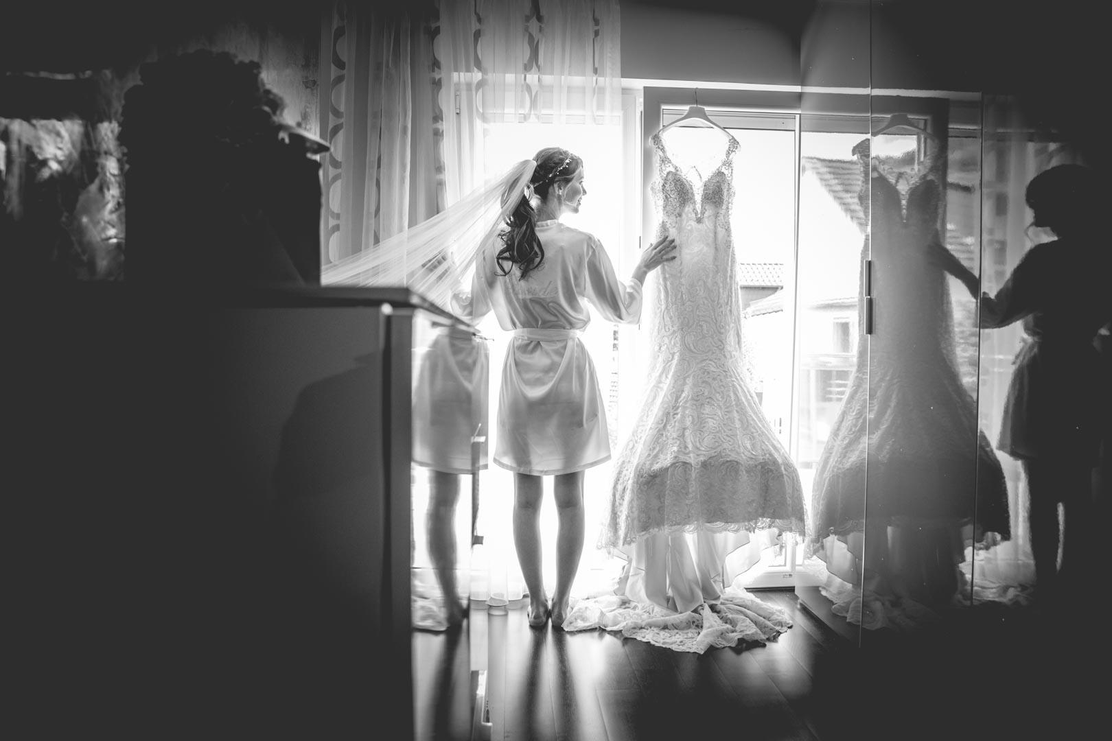 Braut betrachtet im Gegenlicht ihr Hochzeitskleid, das vor dem Fenster hängt